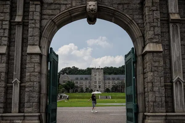 فردی با چتری سفید از میان یک قوس سنگی در محوطه دانشگاه عبور می‌کند.