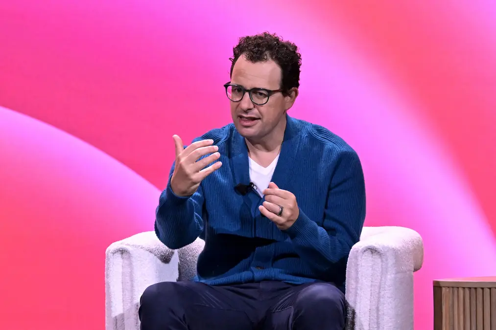 داریو آمودی روی یک صندلی سفید جلوی پسزمینهٔ صورتی مینشیند و با حرکات پرشور صحبت میکند.