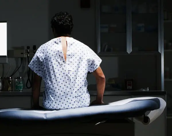 آقایی که از پشت دیده می‌شود، پوشاک بیمارستانی بر تن دارد و در یک اتاق تاریک بر روی میز آبی رنگ نرم‌نشین نشسته است.